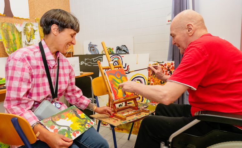 Ein Herr ist mit einer Betreuerin im Atelier und bemalt eine Leinwand